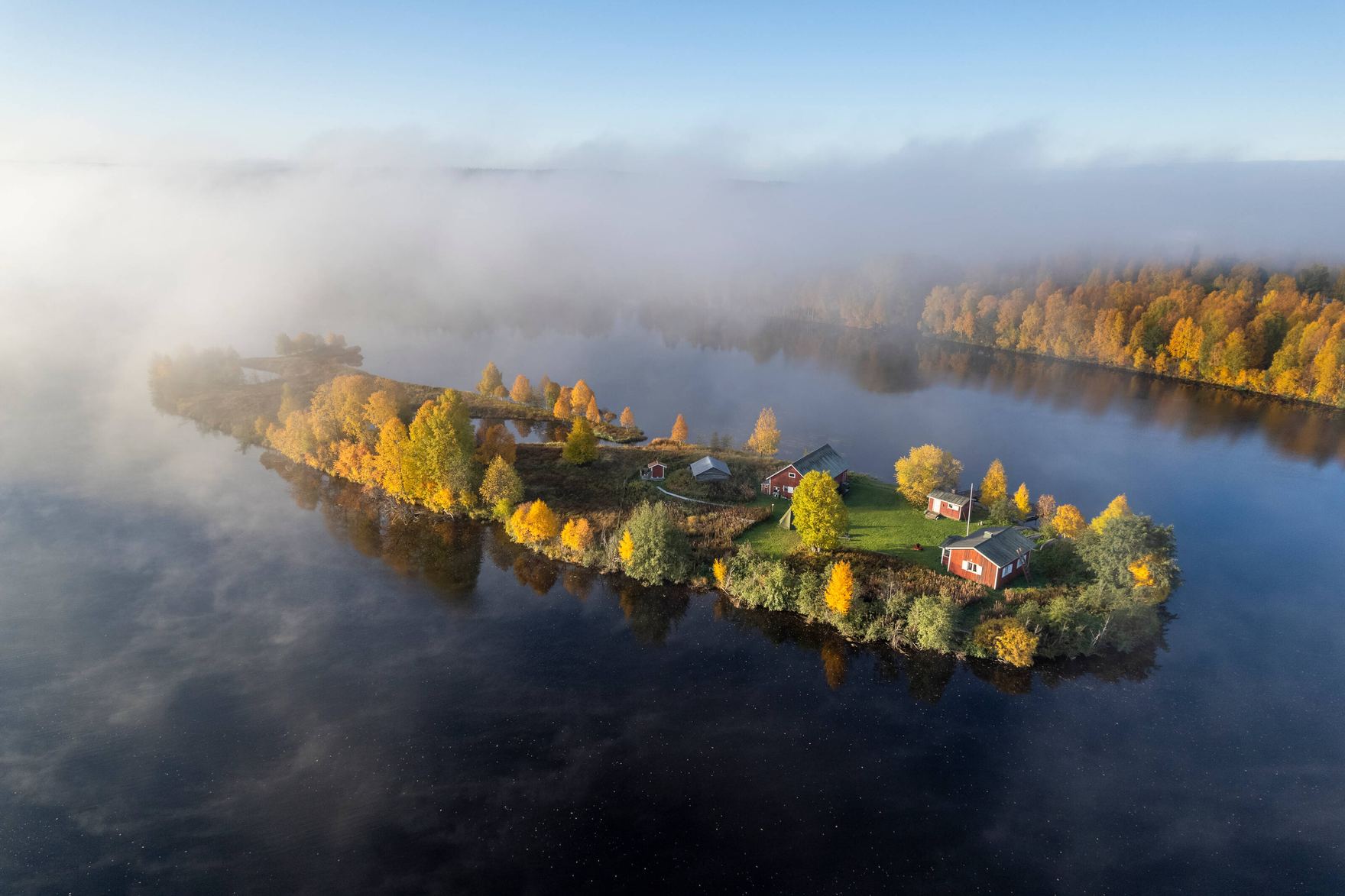 Aerial view of a small forested island with red cabins in a misty autumn lake.